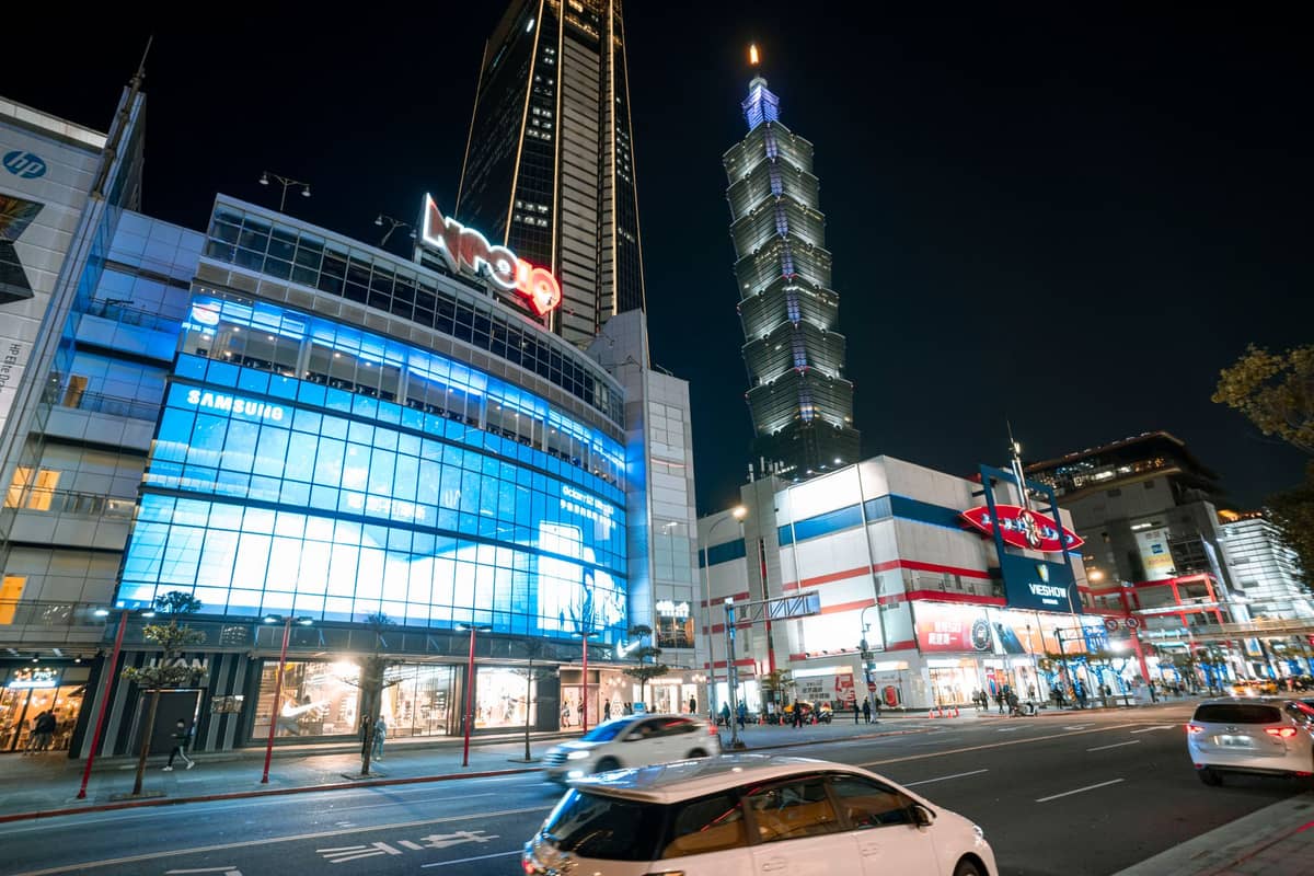 Nächtliche Straßenansicht in Taipei mit hell erleuchteten Gebäuden, pulsierendem Stadtleben und dem beleuchteten Taipei-101-Turm im Hintergrund.