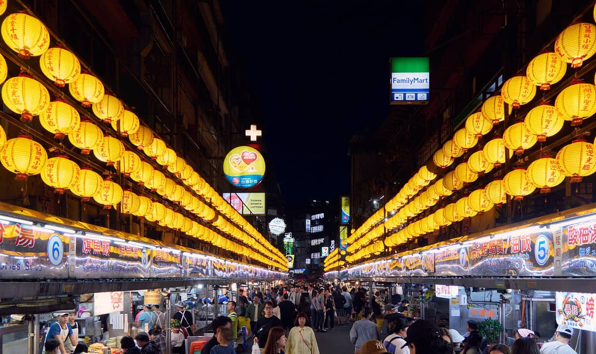 Überfüllter Keelung Miaokou Nachtmarkt bei Nacht mit vielen gelben Laternen, Essensständen auf beiden Seiten und Menschen, die durch die Straße schlendern.