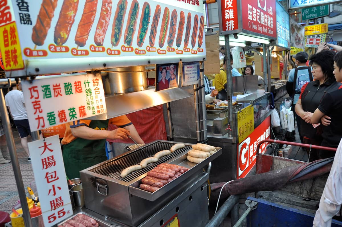 แผงอาหารในตลาดกลางคืนไทเปที่คึกคัก กำลังปิ้งไส้กรอกแบบดั้งเดิม พร้อมชาวบ้านที่รอสั่งอาหารและป้ายภาษาจีนสีสันสดใสด้านบน