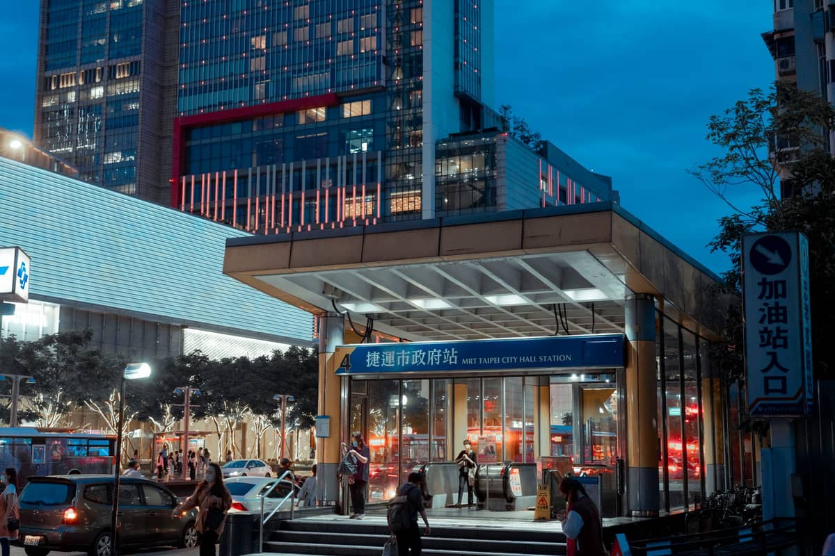 วิวยามเย็นของทางเข้าสถานี Taipei MRT พร้อมผู้คน แสงไฟของเมือง และอาคารสมัยใหม่ในพื้นหลัง