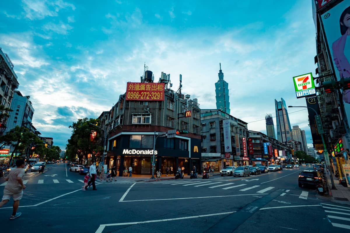 Weitläufige Straße in Taipeh in der Abenddämmerung mit leuchtenden Stadtschildern, einem McDonald's und dem markanten Wolkenkratzer Taipei 101 am Horizont.