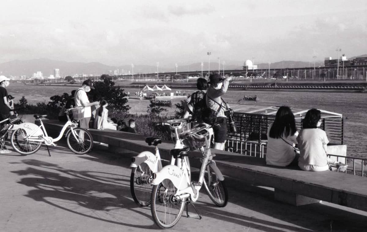 Menschen, die sich in einem Uferpark in Taipei entspannen, mit in der Nähe abgestellten YouBike-Fahrrädern und einer im Hintergrund sichtbaren Brücke.