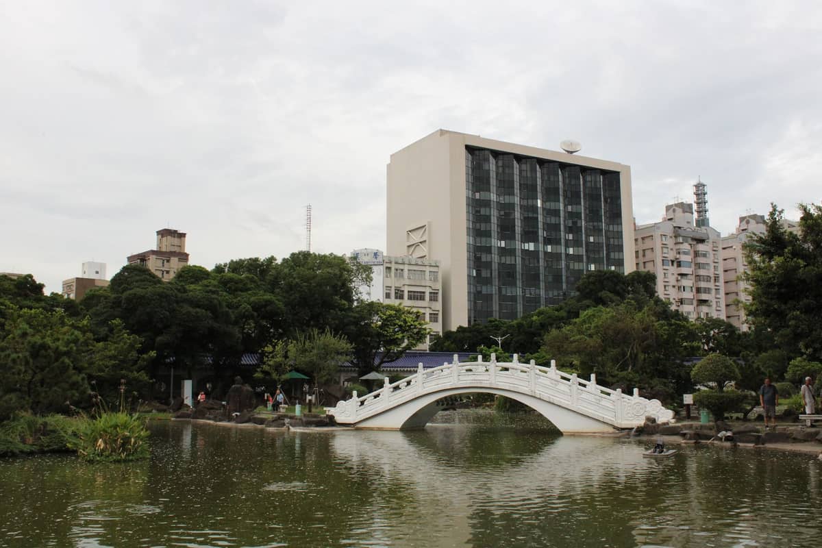 Blick auf einen Stadtpark in Taipeh mit einer traditionellen weißen Bogenbrücke über einen Teich, umgeben von Bäumen und Stadtgebäuden im Hintergrund.