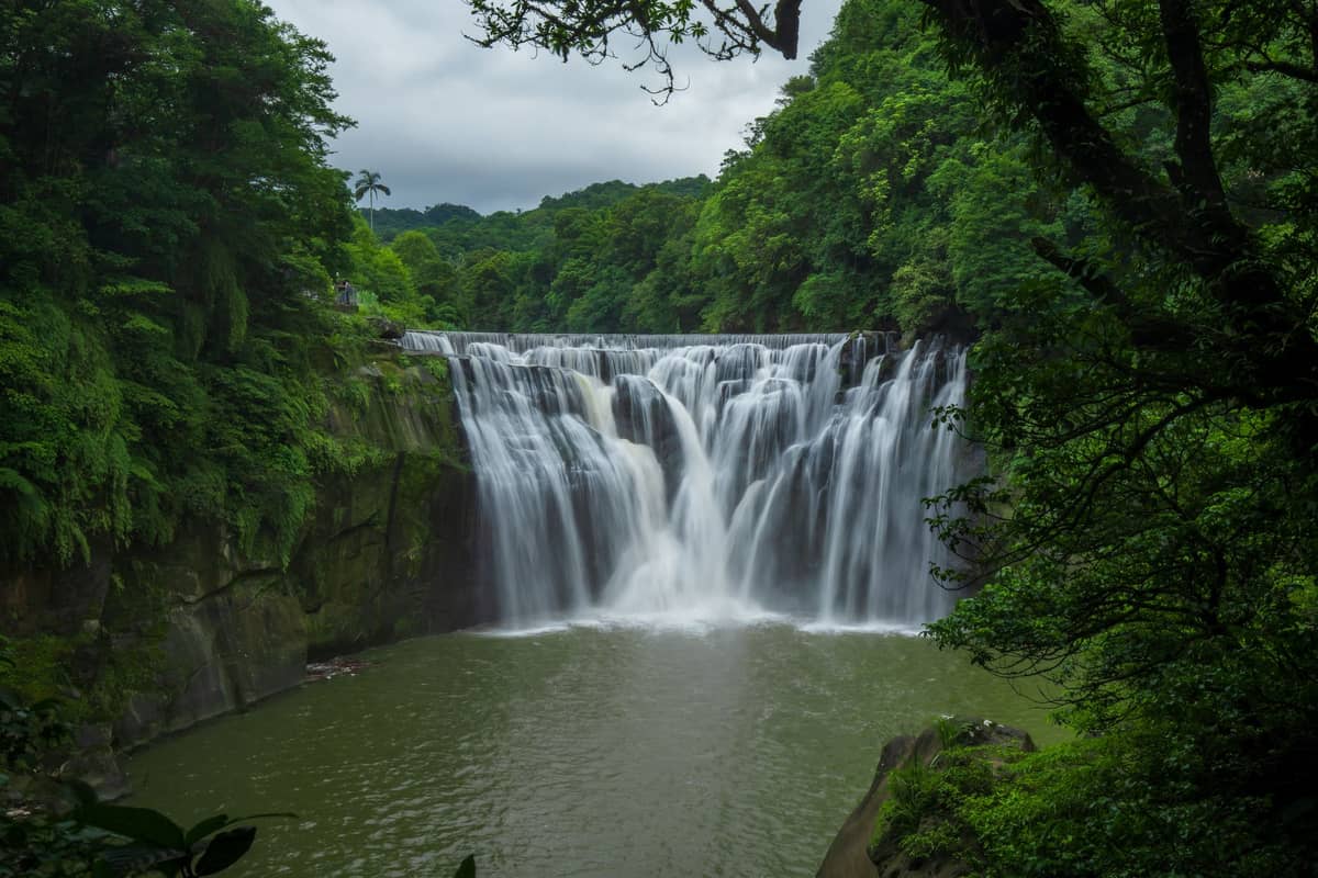 Weitläufiger Blick auf den Shifen Wasserfall, umgeben von üppig grünen Bäumen, mit herabstürzendem Wasser in ein Becken darunter, als beliebtes Naturziel in der Nähe von Taipei.