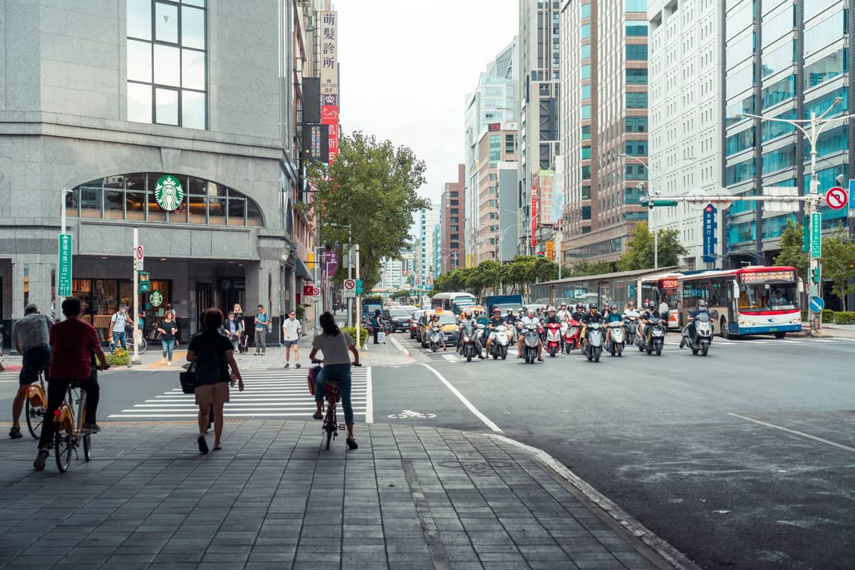 Belebte Straßenszene in Taipeh mit Fußgängern, Radfahrern, Bussen und Rollern an einer Kreuzung, gesäumt von modernen Hochhäusern.