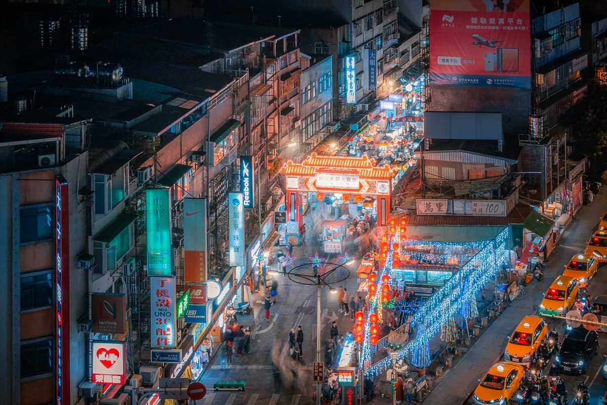 Luftaufnahme des berühmten Raohe Street Nachtmarkts in Taipeh bei Nacht – leuchtende Neonlichter, Essensständer, Menschenmassen und ein rotes Eingangstor.