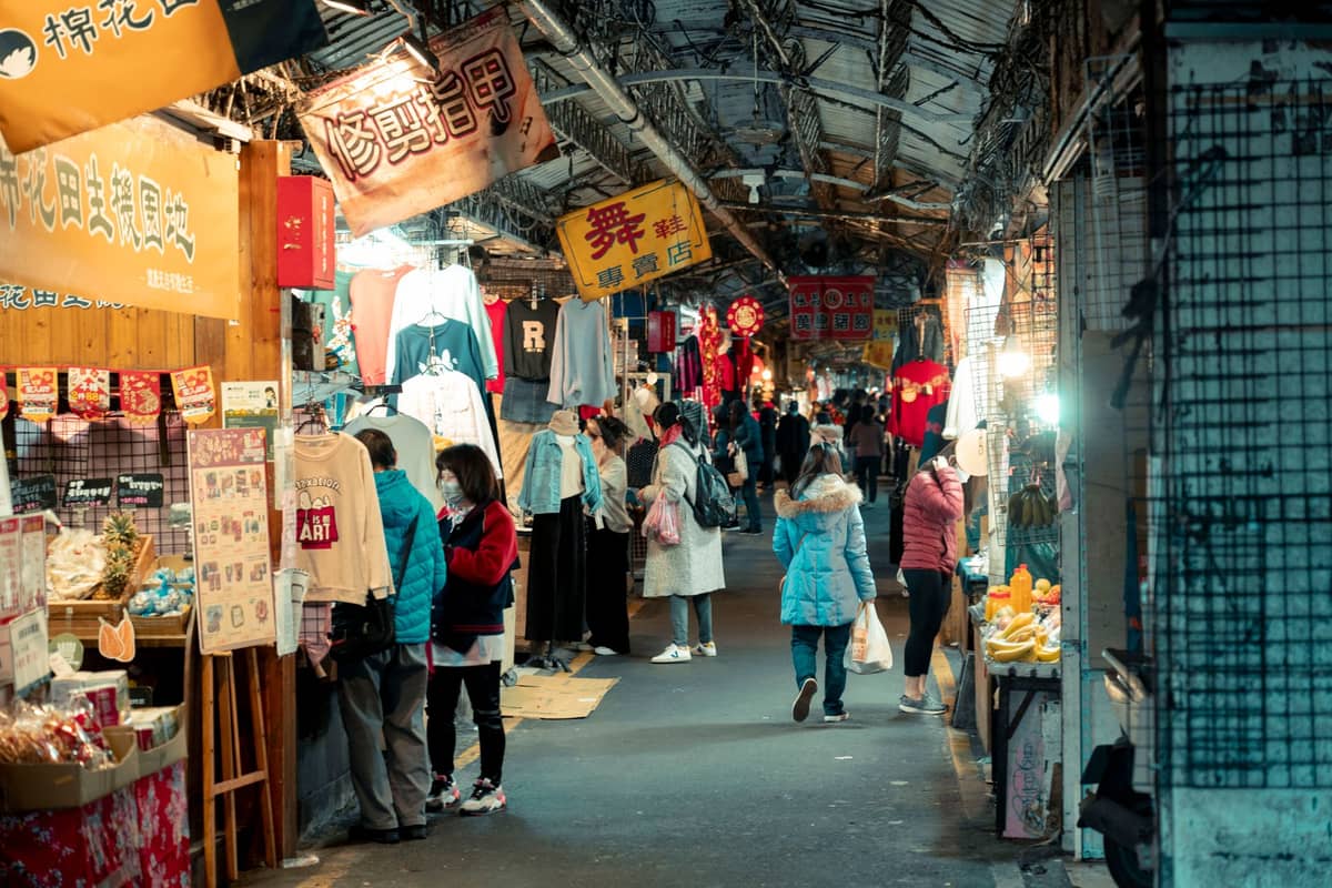 Menschen stöbern an nächtlichen Ständen, die Kleidung, Snacks und Kuriositäten unter hellen Lichtern in einem überdachten Nachtmarkt in Taipei verkaufen.