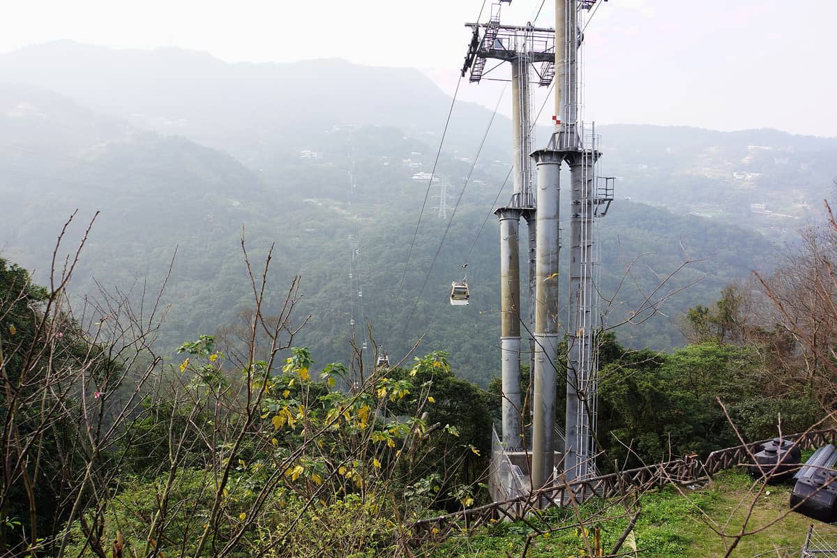Eine lange Seilbahnlinie der Maokong Gondola, die üppige grüne Berge in Taipei überquert, mit Stütztürmen und nebligen Hügeln im Hintergrund.