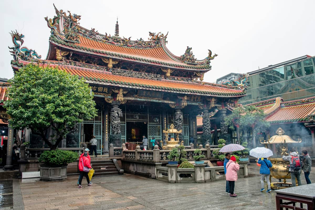 Besucher mit Schirmen und Räucherstäbchen vor der reich verzierten Halle des Longshan-Tempels, einem der bekanntesten Tempel Taipeis.