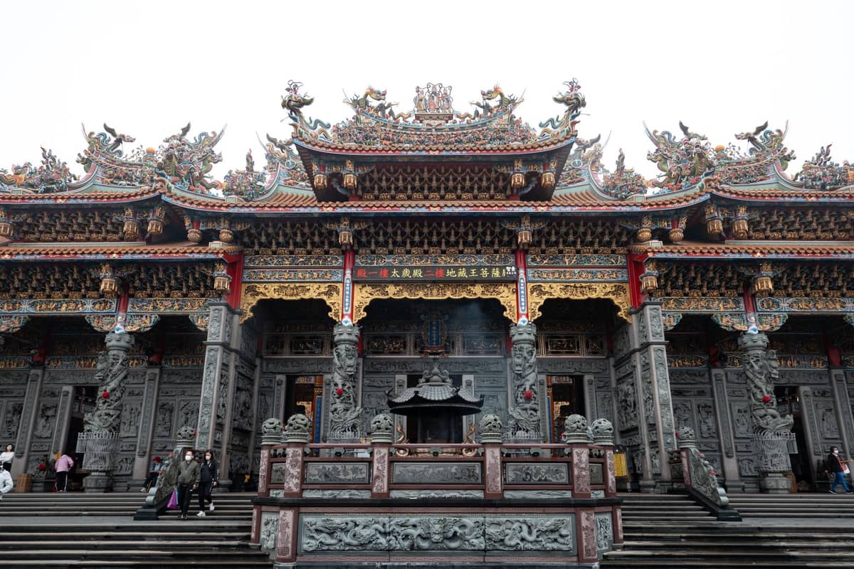 Weitwinkelansicht einer verzierten Tempelfassade mit traditionellem chinesischen Dach und detaillierten Schnitzereien, Menschen, die Steinstufen davor hinaufgehen.