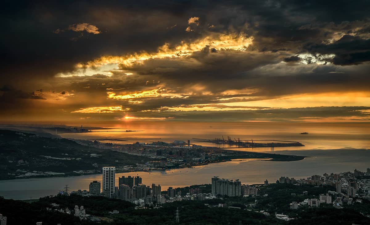 Dramatischer Sonnenuntergang über der Mündung und dem Hafen des Tamsui-Flusses, mit der Uferpromenade und den Hügeln Taipeis im Vordergrund