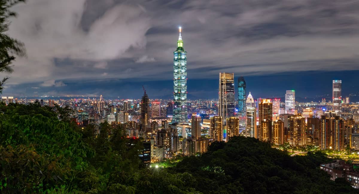Nächtliches Panorama der Skyline von Taipei mit dem beleuchteten Taipei 101-Turm, der sich über die Stadtlichter und grüne Hügel im Vordergrund erhebt.
