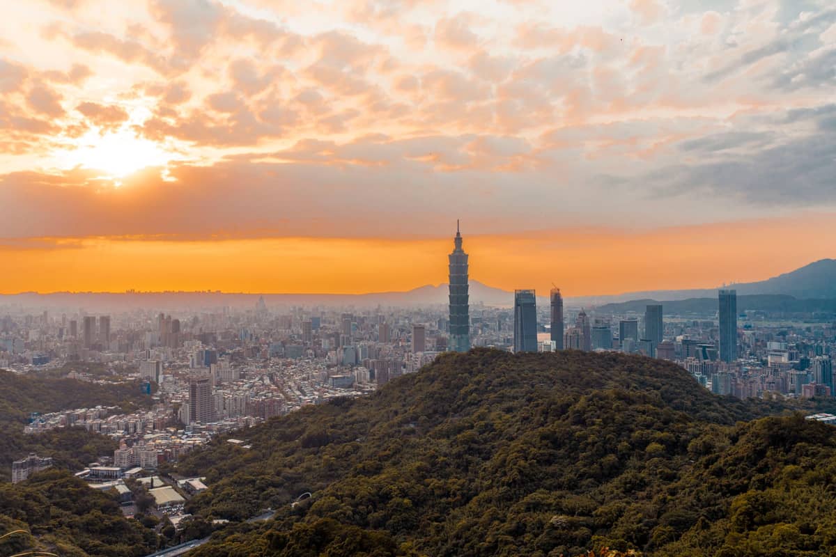 Weiter Blick auf die Skyline von Taipeh bei Sonnenuntergang, mit dem Taipei 101 und umliegenden Wolkenkratzern über üppig grünen Hügeln und dramatisch orangefarbenen Wolken.
