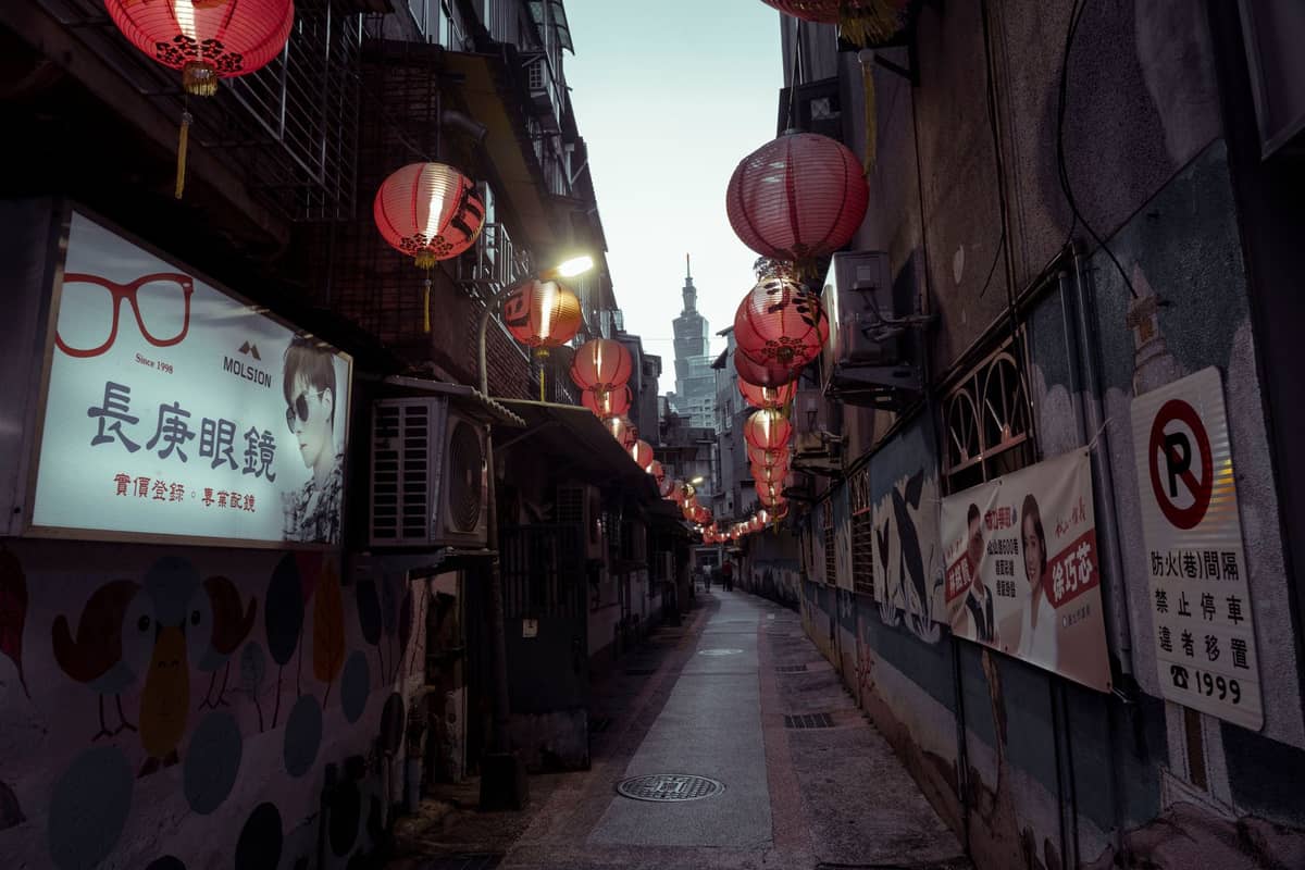 Enge Gasse in Taipei, gesäumt von roten Laternen, Tempelspitze in der Ferne sichtbar, Schilder auf Chinesisch, Atmosphäre einer Stadt zum Flanieren.