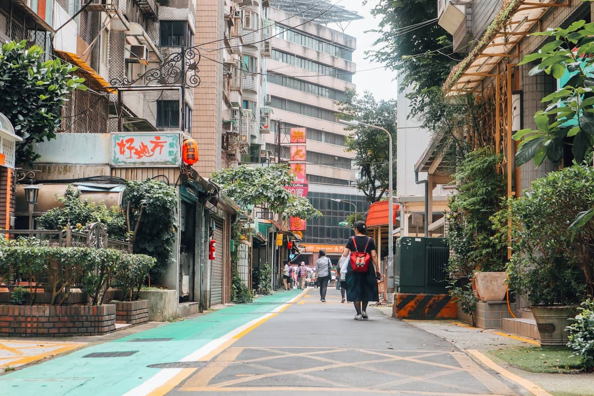 Eine schmale, belebte Gasse in Taipeh mit niedrigen Gebäuden, kleinen Läden und Grünpflanzen, im Hintergrund schlendern Fußgänger entlang.