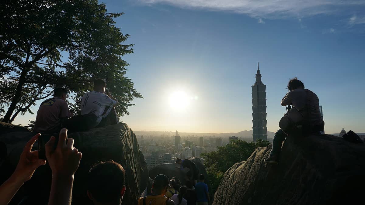 Touristen sitzen bei Sonnenuntergang auf Felsen am Elephant Mountain und blicken auf den Taipei 101 und die Skyline von Taipei.