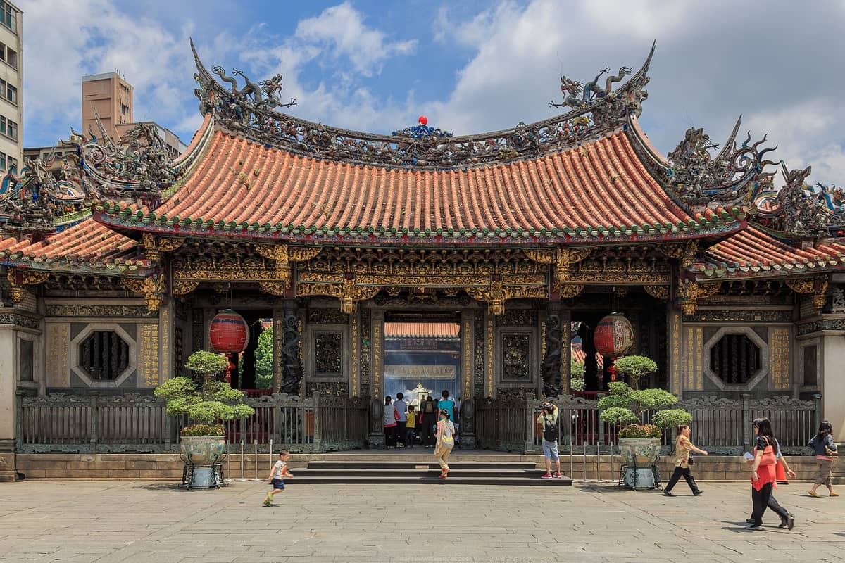 Weitwinkelansicht des Äußeren des Longshan-Tempels in Taipei mit verziertem Dach, detaillierten Schnitzereien, roten Laternen und Besuchern, die davor spazieren.