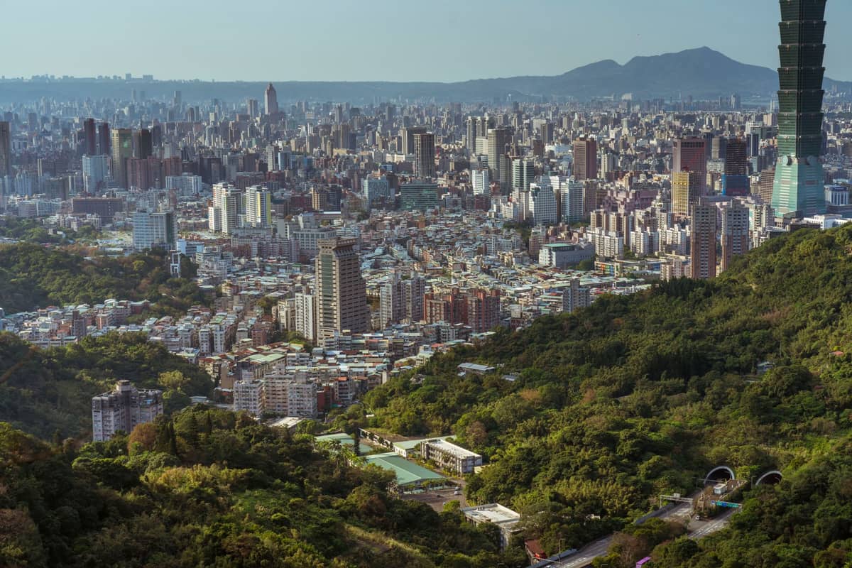 Panoramablick auf die Skyline von Taipeh von oben, mit dem Taipei 101 auf der rechten Seite und bewaldeten grünen Hügeln im Vordergrund.