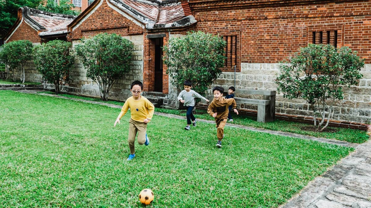 Vier Kinder spielen Fußball auf grünem Rasen vor alten roten Backsteingebäuden in Taipeh – eine familienfreundliche Outdoor-Atmosphäre.