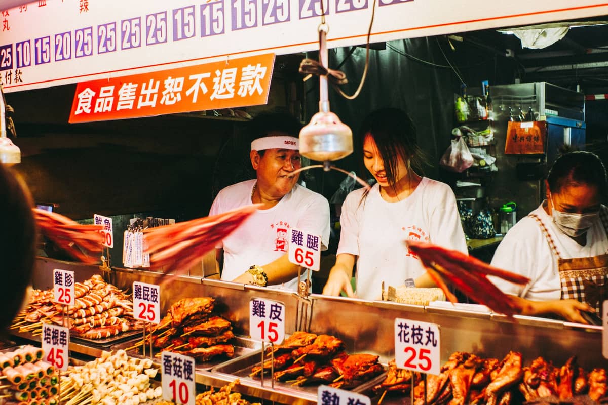 Freundliche Verkäufer hinter einem hell erleuchteten Imbissstand mit Spießen und Snacks auf einem belebten Nachtmarkt in Taipei.
