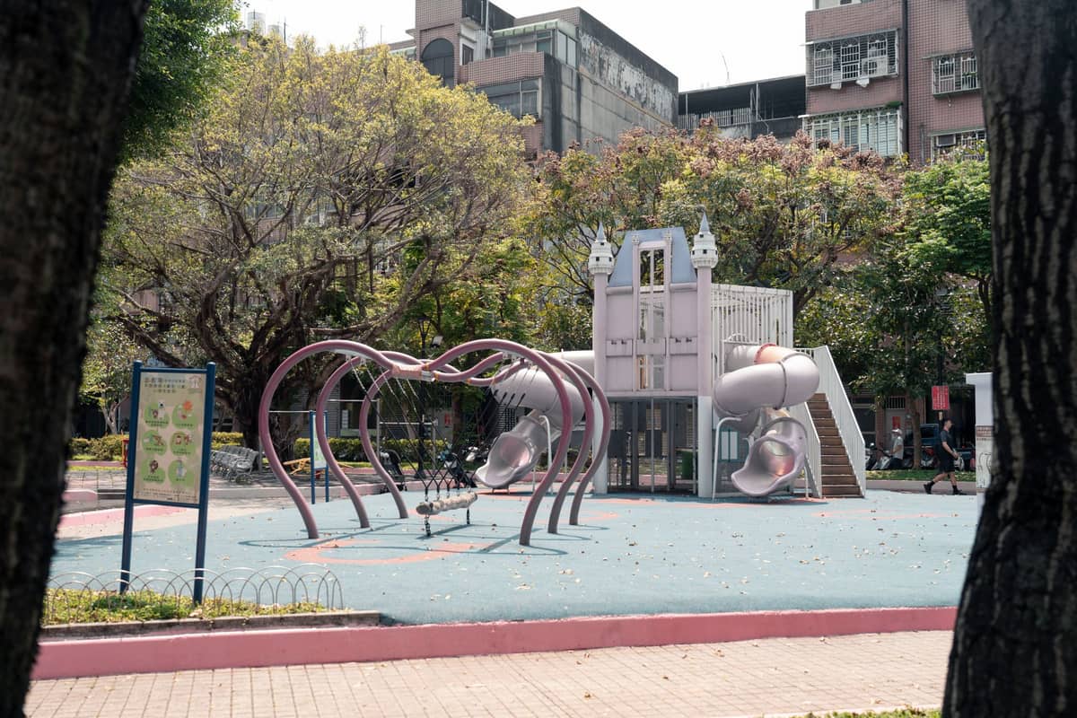 Ein schattiger Spielplatz mit Rutschen und Klettergerüsten in einem grünen Park, umgeben von Bäumen und städtischen Gebäuden.
