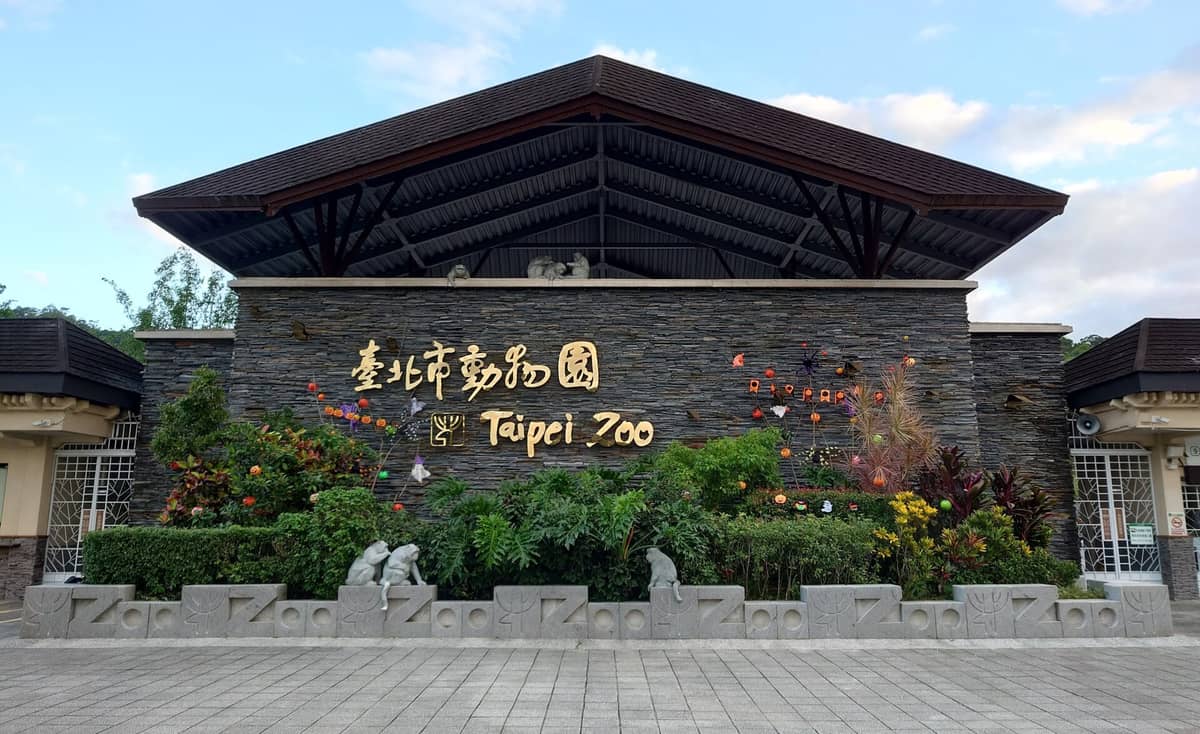 Haupteingang des Taipei Zoos mit großem Schild auf Chinesisch und Englisch, umgeben von Grünpflanzen und Dekorationsblumen unter einem geschwungenen Dach.