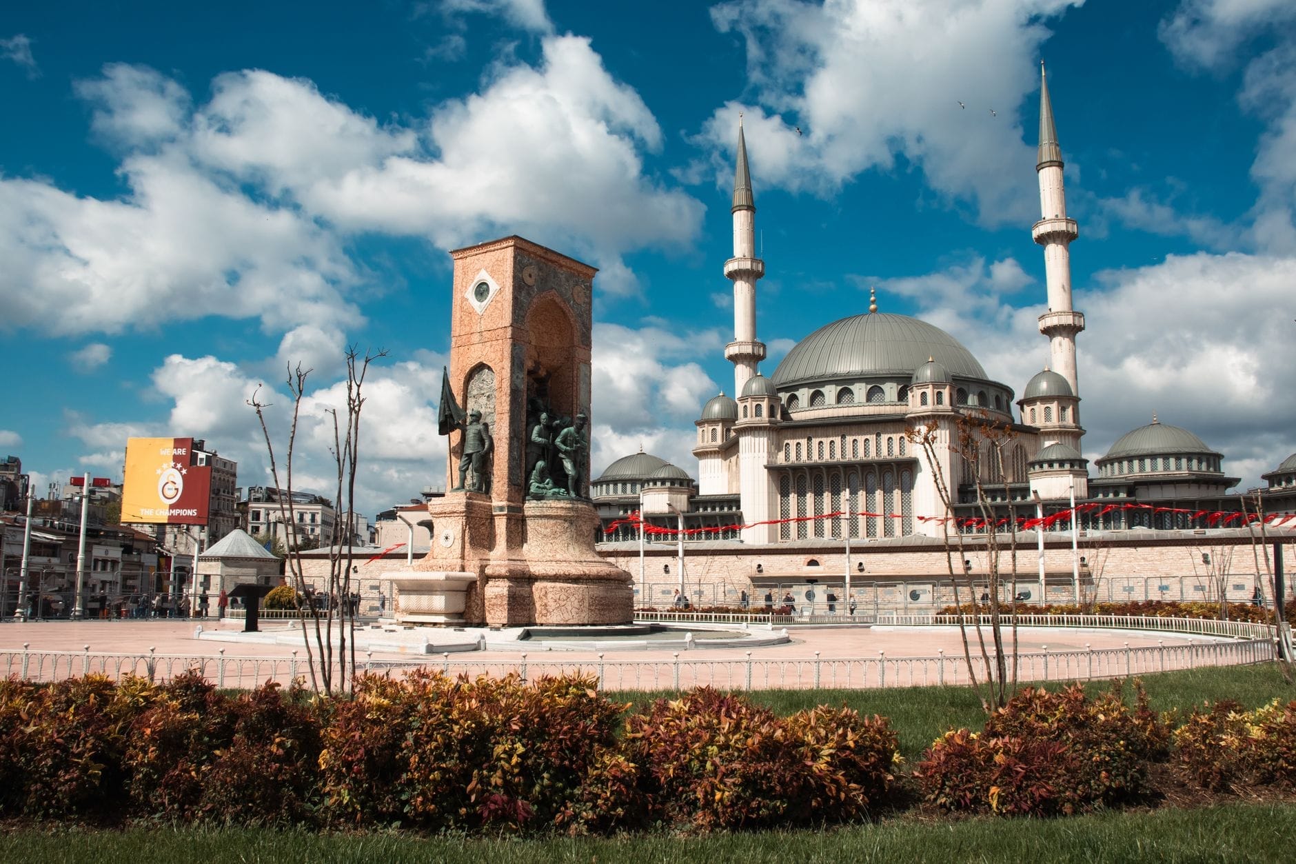 Vista panoramica di Piazza Taksim con il Monumento alla Repubblica al centro e la Moschea di Taksim sullo sfondo, sotto un cielo azzurro con nuvole.