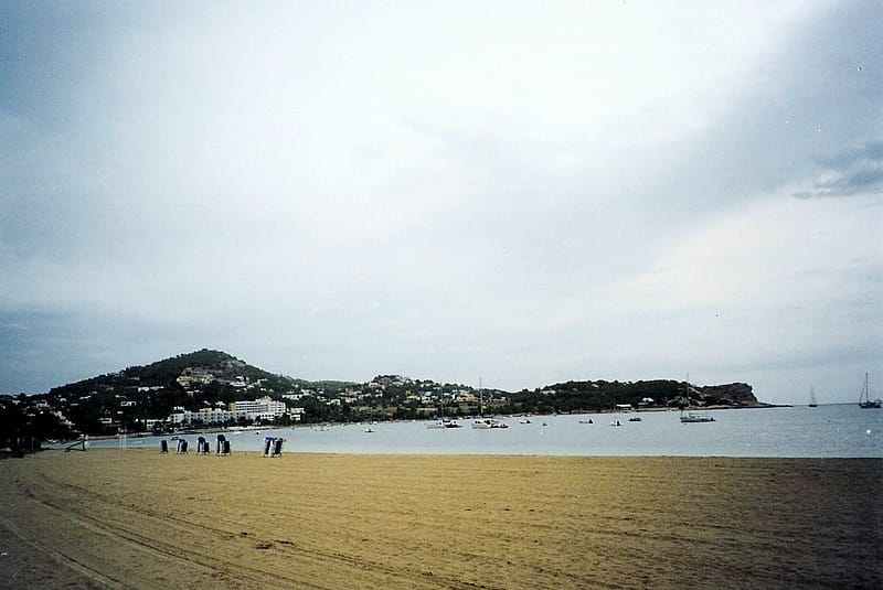 Vista ampia della spiaggia di Talamanca a Ibiza con riva sabbiosa, acque calme, edifici sullo sfondo e barche ancorate vicino alla costa.