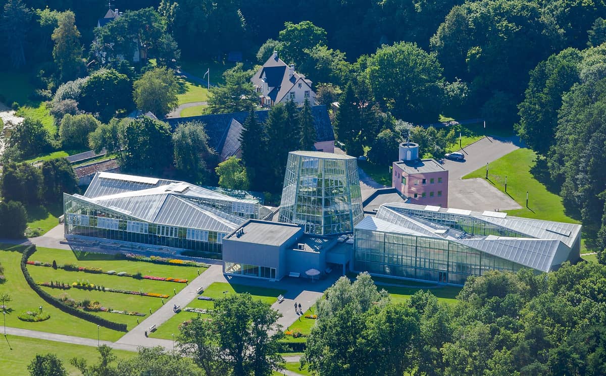 Luftaufnahme des Gewächshauskomplexes des Tallinner Botanischen Gartens, umgeben von Rasenflächen, Gartenbeeten und einem Waldgebiet in Pirita.