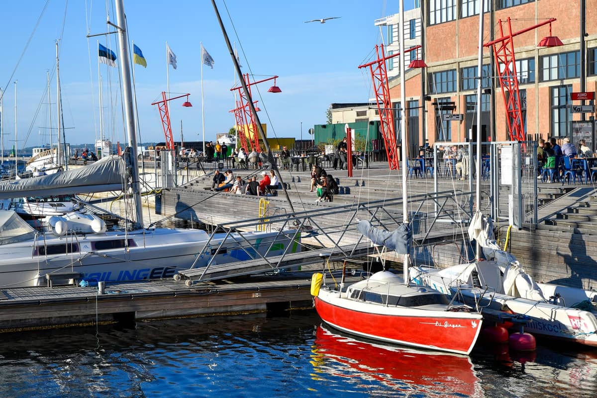 Boote und Yachten, die entlang einer lebhaften Uferpromenade in Tallinn vertäut sind, mit Menschen, die das sonnige Wetter, moderne Gebäude und rote dekorative Laternen genießen.