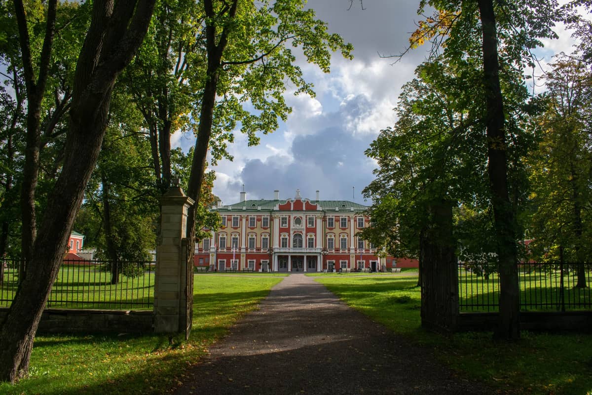 Ein prächtiger Barockpalast, der Kadriorg-Palast, von einem baumgesäumten Weg in seinem grünen Park in Tallinn aus gesehen, unter einem teils bewölkten Himmel.