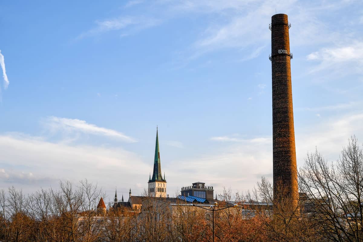 Tallinns Stadtsilhouette mit einem hohen Backsteinschornstein im Kontrast zum grünen Turm der Olaikirche und historischen Dächern, aufgenommen aus der Ferne im warmen Abendlicht.