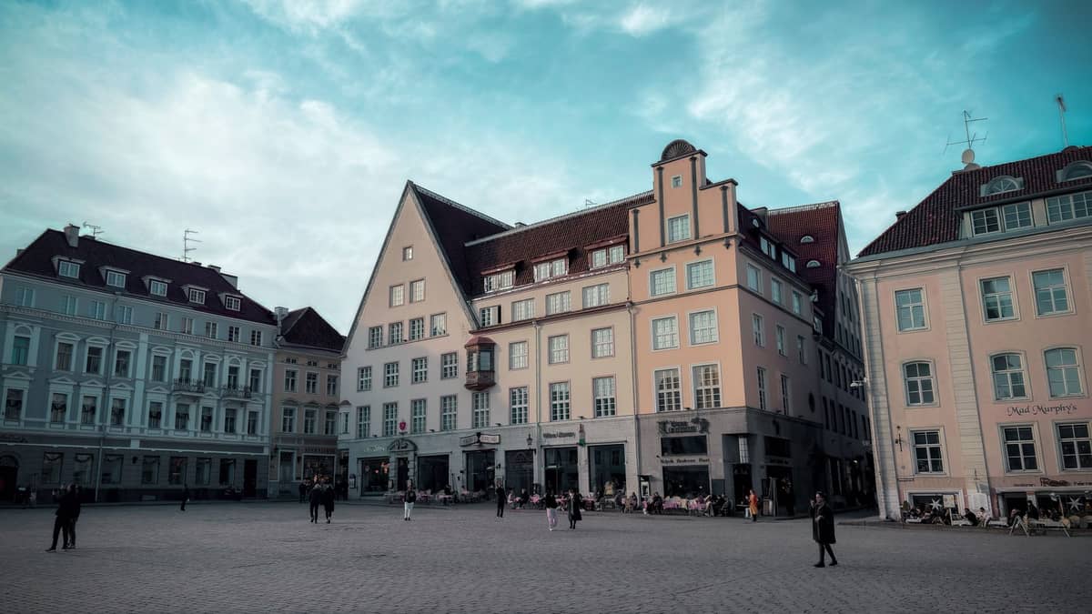 Plac Starego Miasta w Tallinie z pastelowymi średniowiecznymi budynkami, nielicznymi przechodniami, pod błękitnym letnim niebem, pokazujący, że teren nie jest nadmiernie zatłoczony.