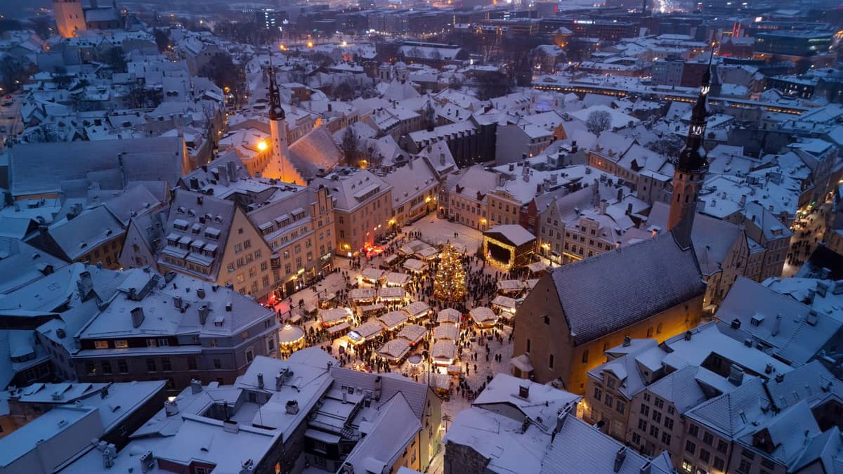Luftaufnahme der verschneiten Tallinner Altstadt in der Dämmerung, mit festlichen Marktlichtern und umliegenden historischen Gebäuden.