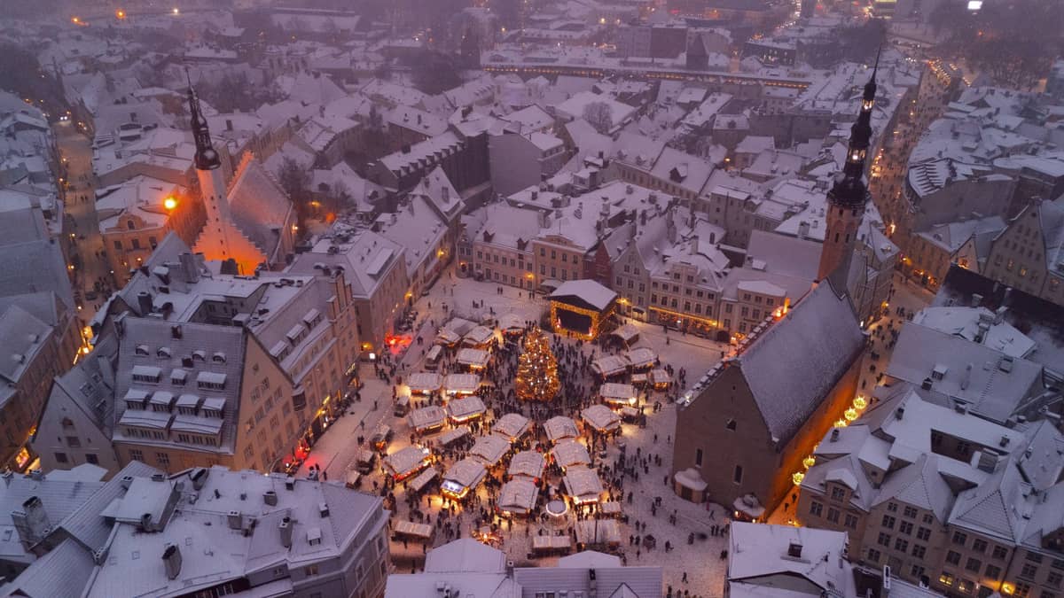 Luftaufnahme der Tallinner Altstadt im Winter mit historischen Gebäuden und einem Weihnachtsmarkt auf dem verschneiten Rathausplatz in der Abenddämmerung.