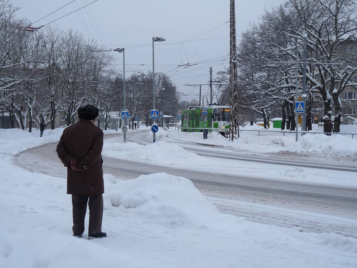 Verschneite Stadtstraße in Tallinn mit einer herannahenden grünen Straßenbahn, wartenden Fußgängern und mit frischem Schnee bedeckten Bäumen.