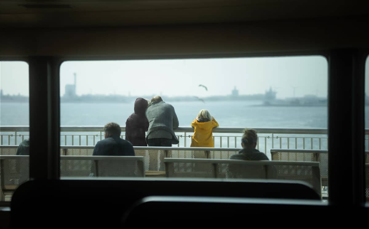 Persone sul ponte di un traghetto che guardano attraverso la baia di Tallinn verso una riva lontana con il porto e lo skyline della città visibili.