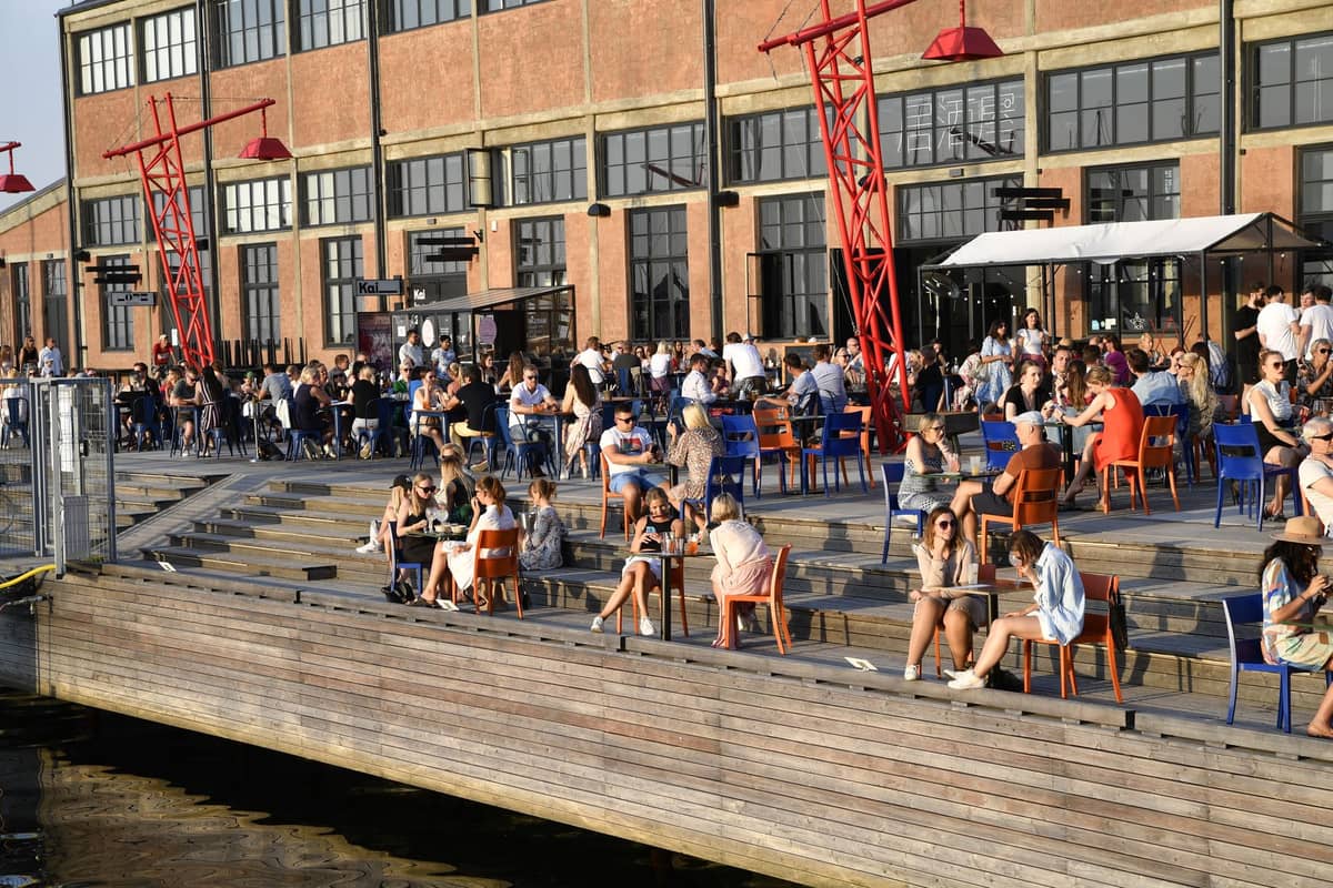 Menschenmengen sitzen und unterhalten sich auf den Terrassen der Noblessner-Uferpromenade in Tallinn im Sommer, mit modernen Backsteingebäuden und roten Kränen im Hintergrund.