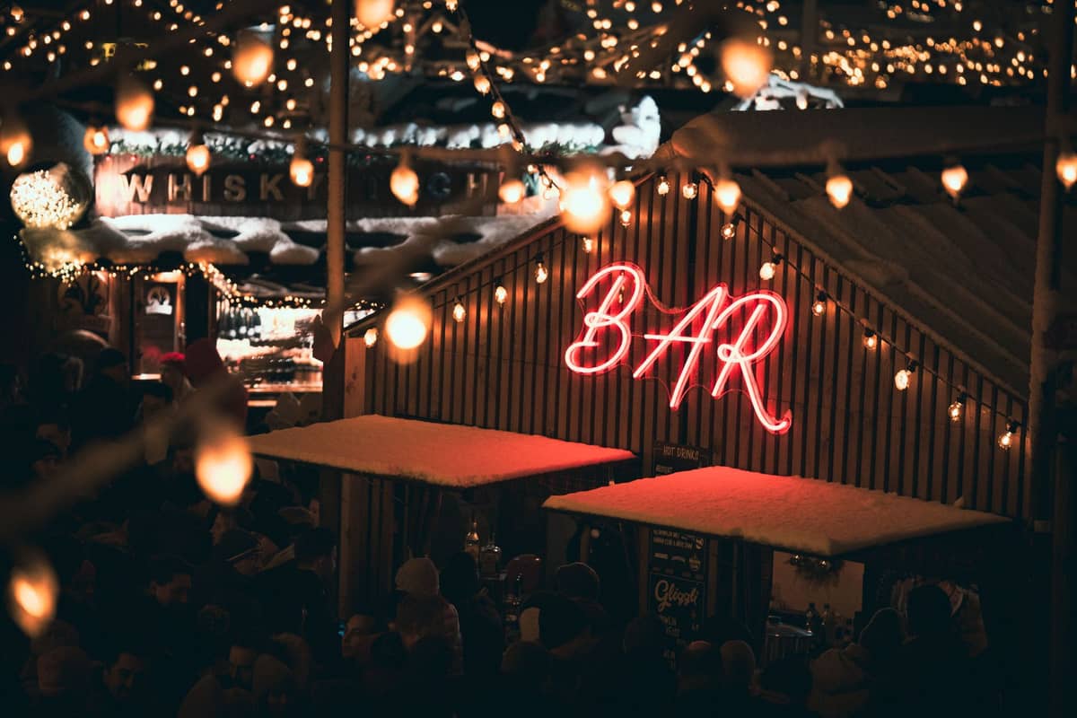 Eine lebhafte Bar im Freien bei Nacht in Tallinn mit leuchtendem Neon-'BAR'-Schild, Menschenmassen und Wintermarktlichtern.