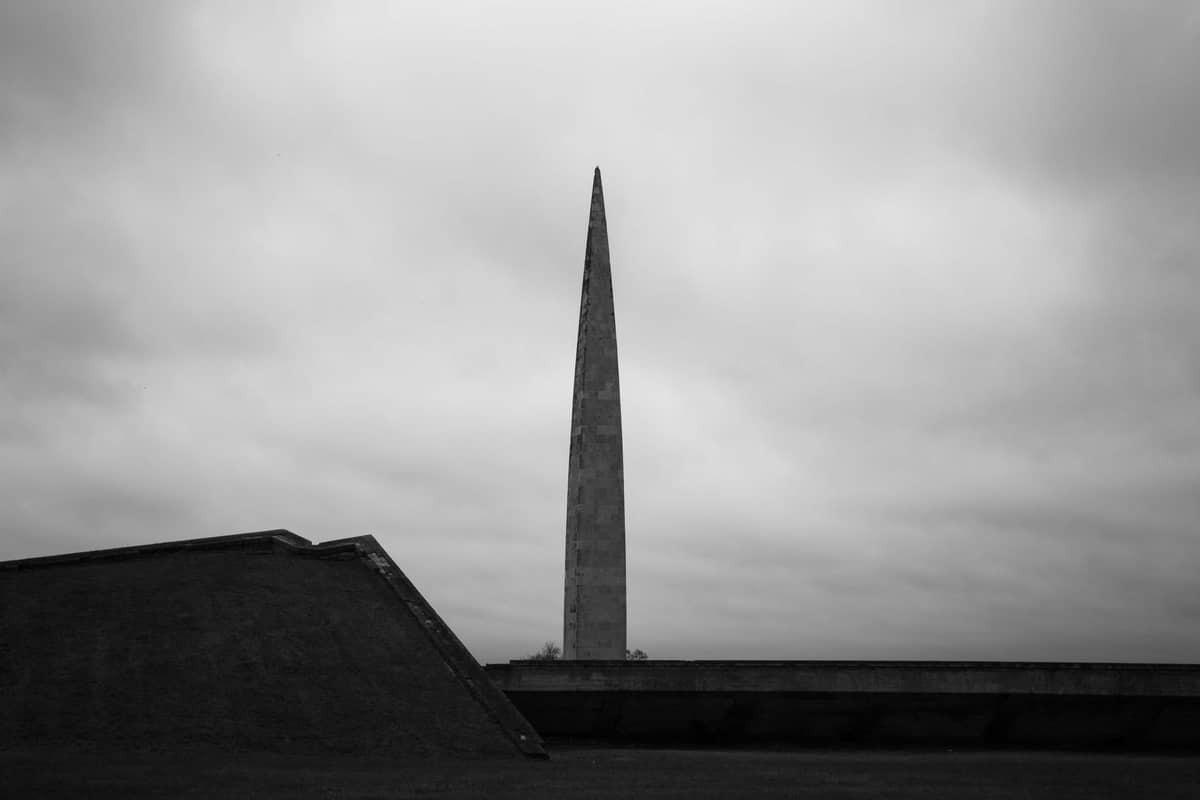 Wysoki, wąski betonowy obelisk z czasów sowieckich wznoszący się na tle pochmurnego nieba, z geometrycznymi formami upamiętniającymi na pierwszym planie w Kompleksie Memorialnym Maarjamäe, Tallin.