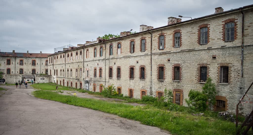 Szeroki widok na zewnętrze więzienia Patarei w Tallinie, ukazujący zwietrzałe kamienne mury, okna z kratami i dziedziniec z kilkoma odwiedzającymi.