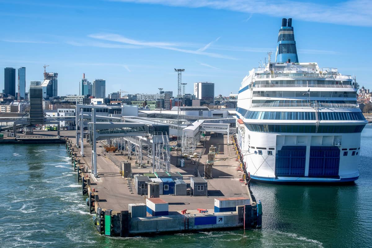 Eine große weiße Fähre liegt an einem modernen Terminal im Hafen von Helsinki, im Hintergrund die Stadtsilhouette und ein klarer blauer Himmel.