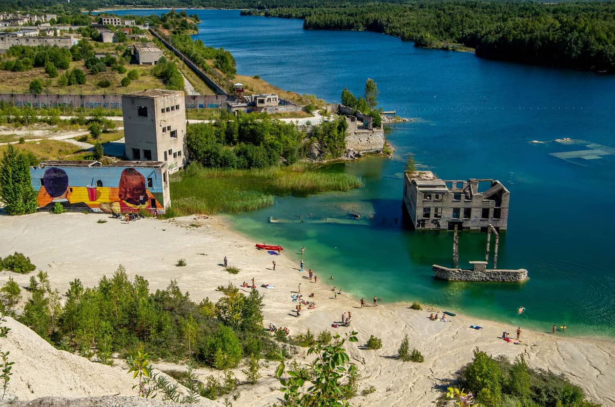 Luftaufnahme des Rummu-Steinbruchs mit türkisblauem Wasser, Sandstrand, Schwimmern, Ruinen, Grünflächen und Graffitiwand an einem sonnigen Tag in der Nähe von Tallinn.