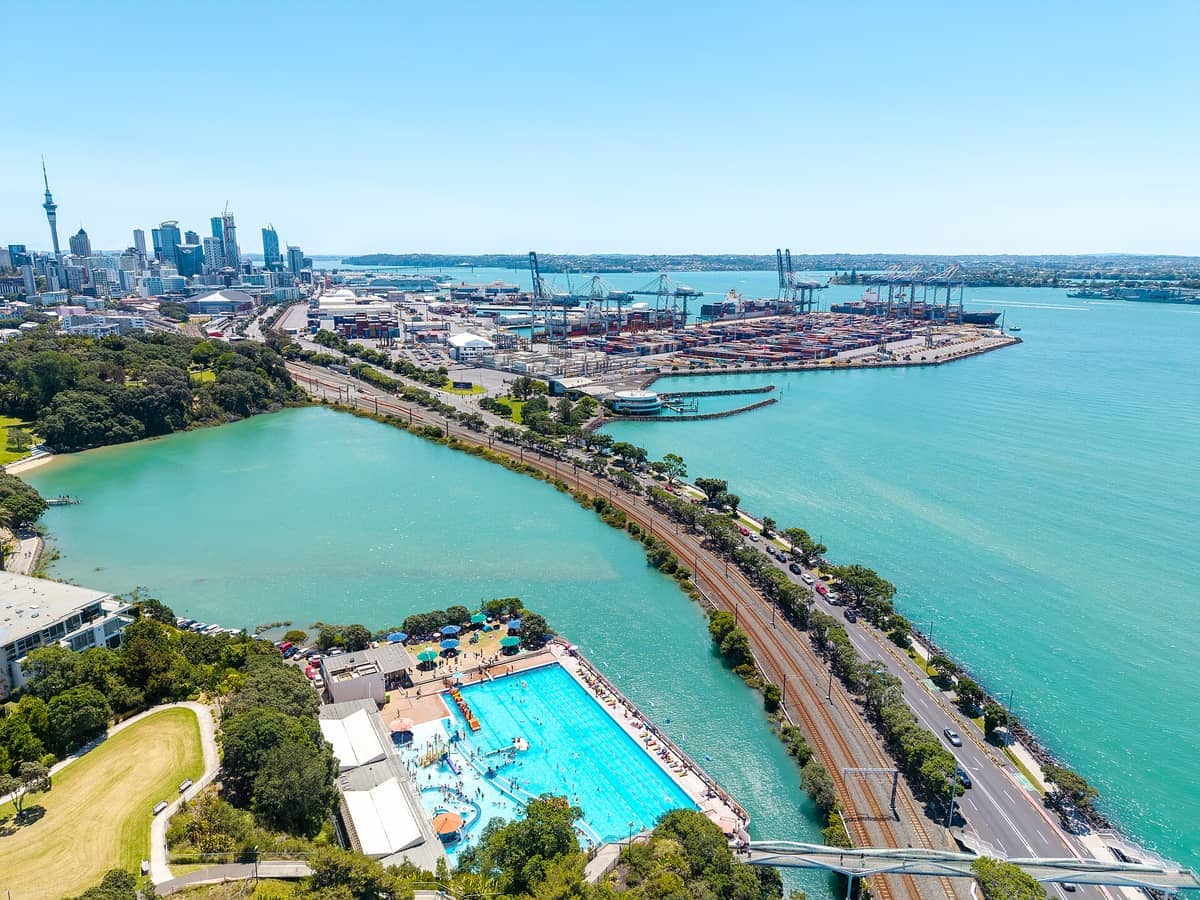 Luftaufnahme des Tāmaki Drive mit funkelndem türkisfarbenem Wasser, Stadtgebäuden, einem belebten Hafen, Schwimmbad und üppigem Grün unter klarem blauem Himmel.