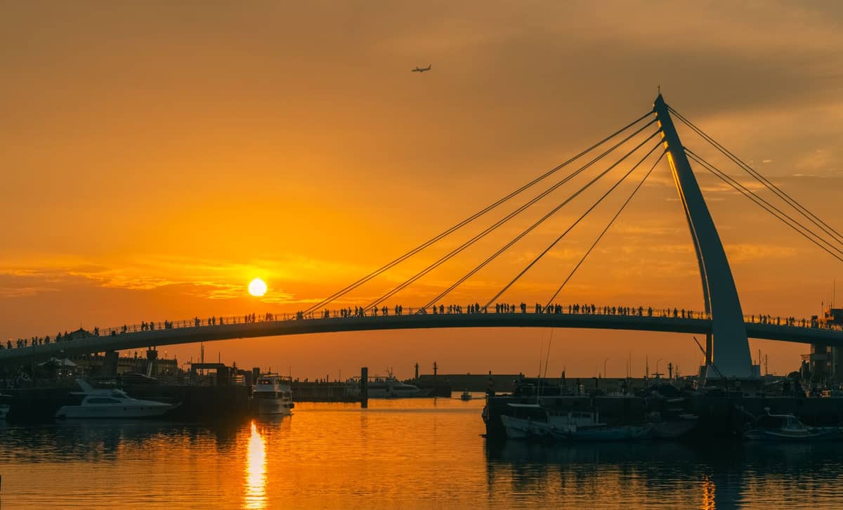 Die Lover's Bridge am Tamsui Fisherman's Wharf als Silhouette vor einem leuchtenden Sonnenuntergangshimmel, darunter Menschen und Boote im Hafen.