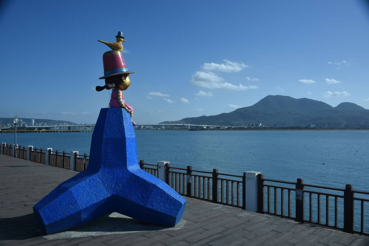Blaue Statue und Uferpromenade mit Blick auf den Tamsui-Fluss, im Hintergrund der Guanyin-Berg unter klarem Himmel am Tamsui Old Street Waterfront.