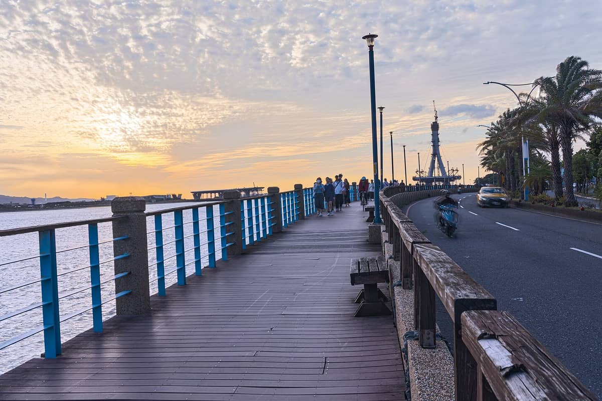 Die Uferpromenade von Tamsui im Sonnenuntergang: Menschen spazieren auf dem Holzweg, im Hintergrund ist die Lover's Bridge und das Flussufer zu sehen.