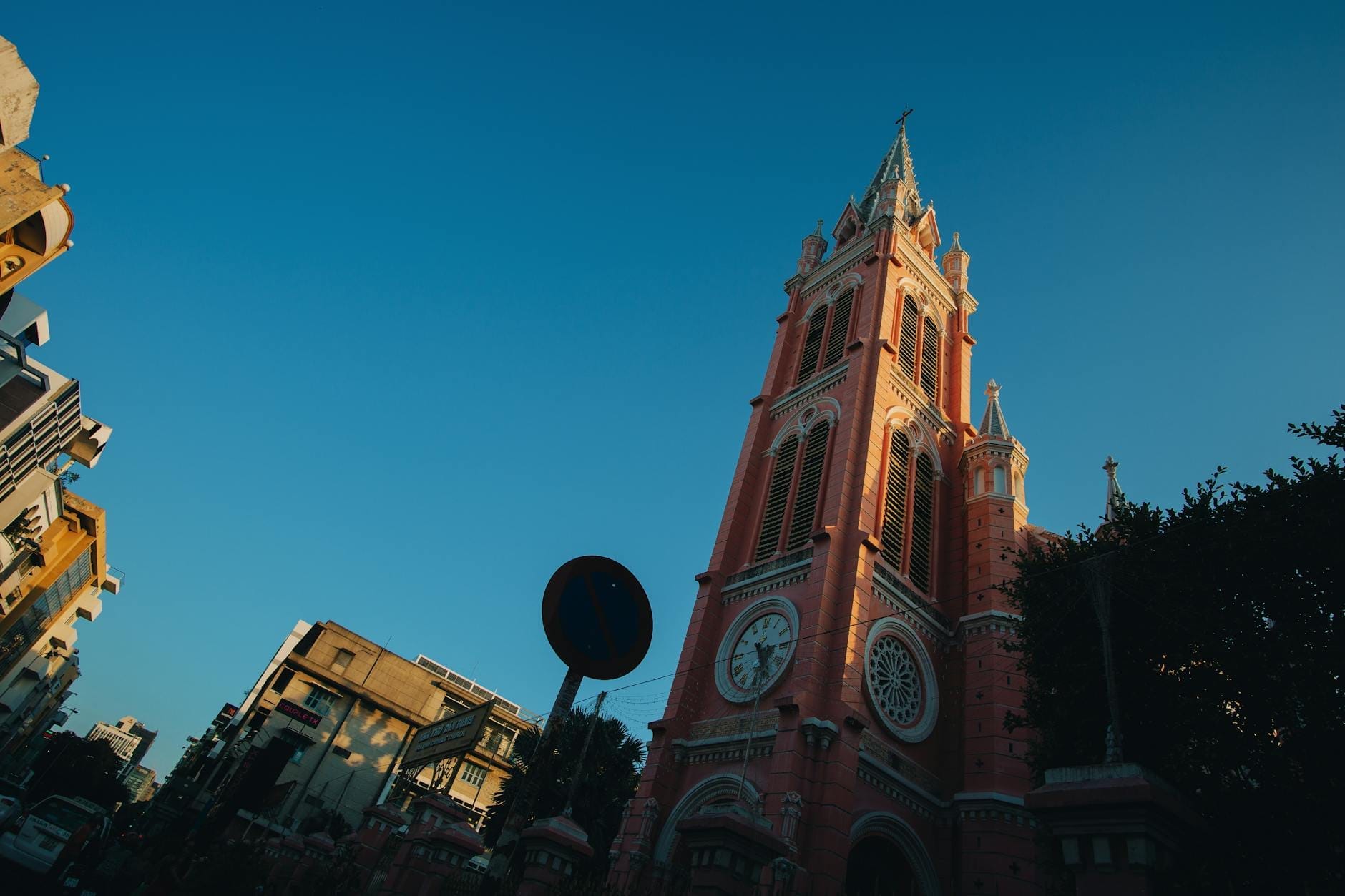 El alto campanario rosado de estilo gótico-románico de la iglesia Tan Dinh se eleva dramáticamente hacia un cielo azul despejado, rodeado de edificios y vegetación en Ciudad Ho Chi Minh.