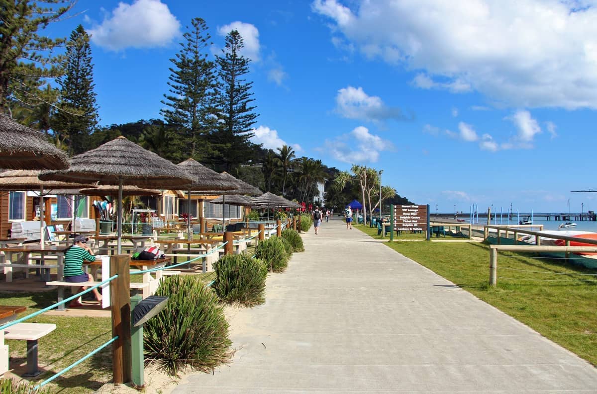 Schattige Picknicktische und Resort-Hütten säumen den sandigen Weg im Tangalooma Island Resort, mit hohen Kiefern und strahlend blauem Himmel darüber.