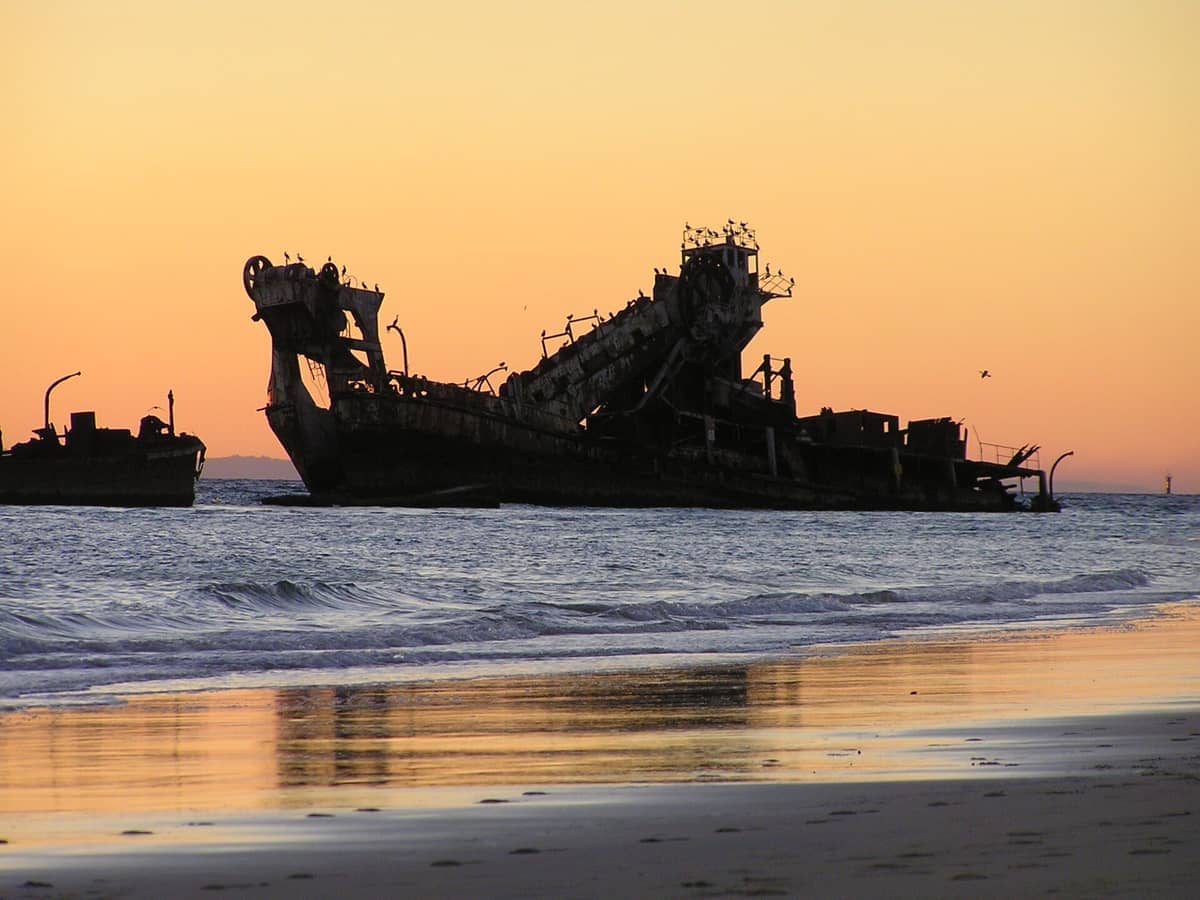 Silhouetten der Tangalooma Wrecks auf Moreton Island spiegeln sich bei Sonnenuntergang in ruhigen Küstengewässern wider, mit Vögeln auf den Strukturen.